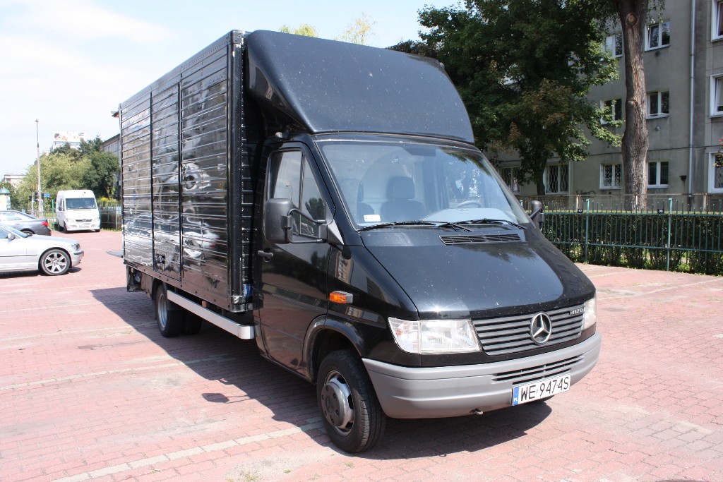 Czarny samochód dostawczy Mercedes-Benz Sprinter z wysoką zabudową, zaparkowany na czerwonej kostce brukowej, widoczny numer rejestracyjny WE 9474S.